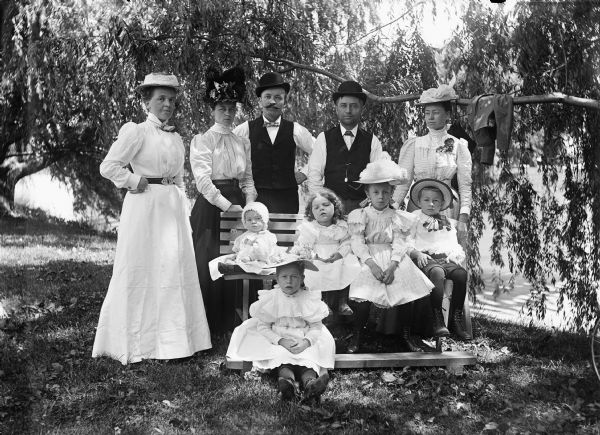 A group of adults and children pose for a portrait at a lakeside setting. Four children are sitting on a bench, and a young girl sits on the ground. In the background is a shoreline with trees.