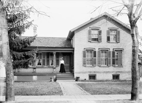 Woman in dress and hat standing on front porch of brick house.