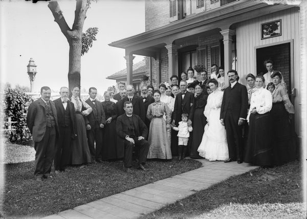 Outdoor group portrait of adults and one child posed in yard in front of a house, with a member of the clergy in the front. Perhaps a wedding portrait.