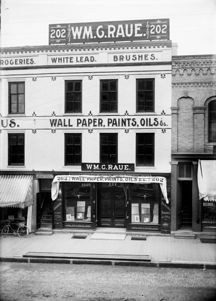 View from across street of storefront of Wm. C. Raue. at 202 Main Street. Signs on the building say: "Wallpaper, Paints, Oils."