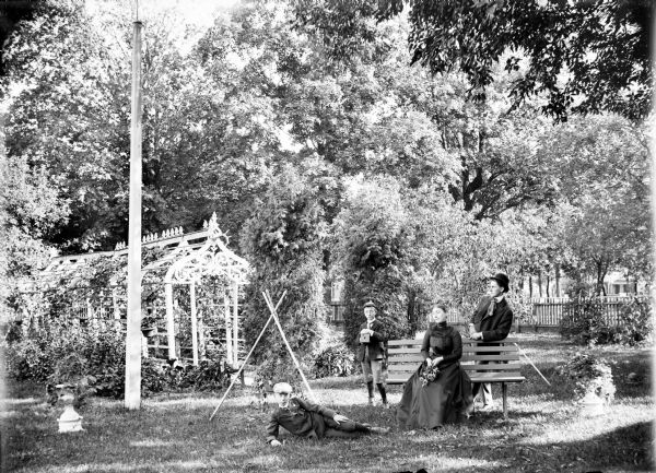 Woman seated on a park bench holding flowers. A young man stands behind her leaning on the bench, and two boys are posed nearby. One boy holds a box camera. An arbor with vines is behind a tall pole on the left.