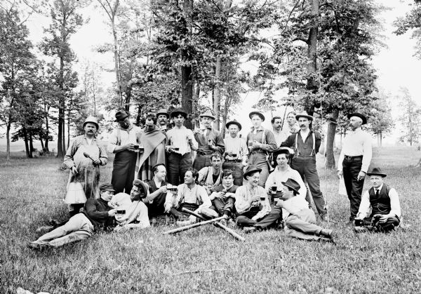 Humorous portrait of large group of men drinking beer. They are in an open, grassy area, with trees behind them. Two baseball bats and a glove are positioned in front of group.