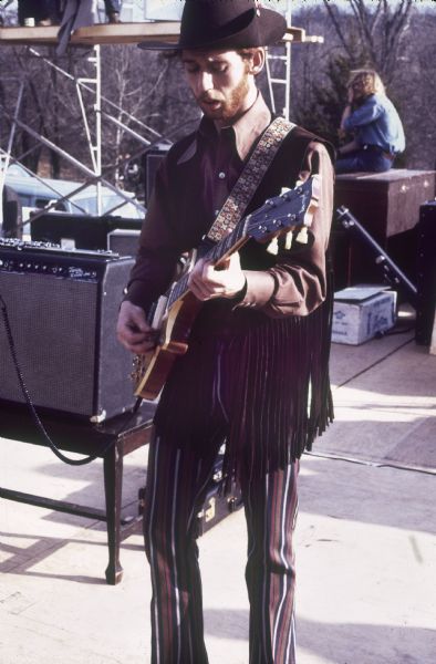 The guitarist for Baby Huey & the Babysitters, possibly Danny O'Neil, stands on stage playing a Gibson Les Paul guitar, wearing a wide brimmed hat and large collared shirt with a fringed vest. Behind him is a Fender Twin Reverb "Black Face" amplifier.