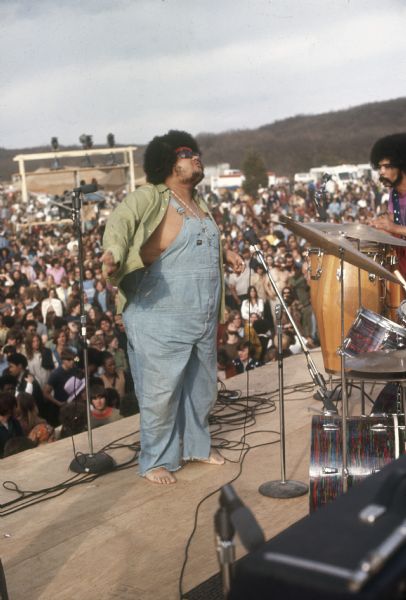 Chicago R&B singer Baby Huey standing barefoot on stage wearing an open green shirt on top of coveralls. A conga player, possibly Plato Jones, is visible on the right side of the photograph. A large crowd is in the background.