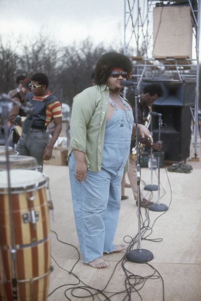 Chicago R&B singer Baby Huey standing barefoot on stage wearing an open green shirt on top of coveralls. Members of his horn section are visible in the background, as well as the guitarist who is possibly Danny O'Neil. It is likely that the tenor saxophone is being played by Byron Watkins and the trumpet by Rick Marcotte.