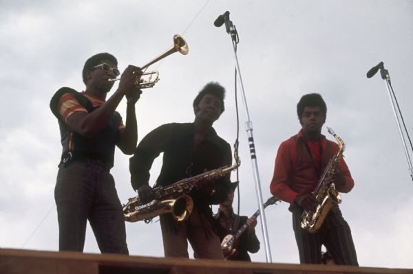 Low angle view of three members of the Baby Huey & the Babysitters horn section, which features a modified trumpet, and a tenor and an alto saxophone. They men are standing on the edge of the stage playing into microphones. They are Turk Littles on trumpet, Charlie Clark on tenor saxophone, and Bobby Baker on alto saxophone.