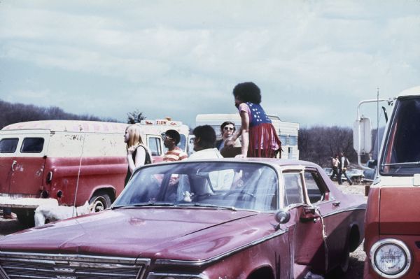 Chicago R&B singer Baby Huey leaning against a car with his trumpeter, (likely Rick Marcotte) and conga player, (possibly Plato Jones) who is wearing an American flag print fringed vest, as well as two or three unidentified people with him. The car they are leaning on has a large dent in the drivers side door, and a white dog can be seen on the left side of the photograph near a van.