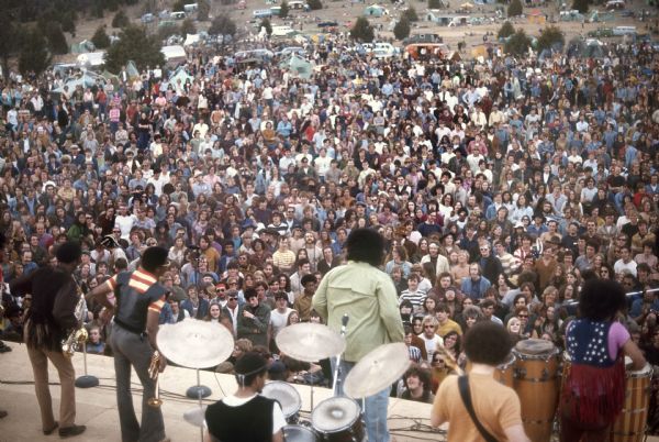 View from above and behind the stage of Chicago R&B act Baby Huey & the Babysitters mid-performance, while the large festival audience stands around the stage. In the far background cars are parked, and tents are set up on a hill.