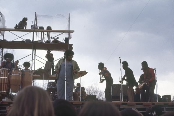 Chicago R&B act Baby Huey mid-performance viewed from the audience. The band was made up of a three man horn section, electric guitar, drums, congas, electric bass, and the singer James Ramey, better known as "Baby Huey." In the background a photographer is standing on a scaffolding behind the stage.
