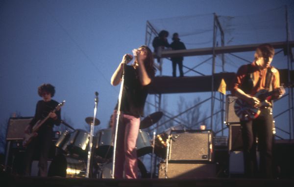 Minneapolis rock band Crow, featuring front man Dave Wagner, guitarist Dick Wiegand, bassist Larry Wiegand, keyboardist Kink Middlemist (not pictured here) and drummer Denny Craswell, on stage at Sound Storm. Two people are visible on the scaffolding behind the stage.