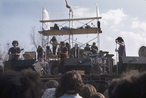 The Grateful Dead mid-performance as viewed from the audience. People are also watching from a scaffolding behind the stage.