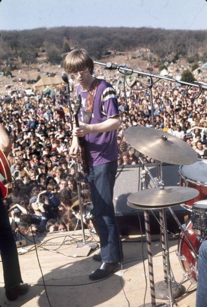 Phil Lesh, bassist for the Grateful Dead on stage wearing a t-shirt reading "Fillmore West." Mickey Hart's drum kit can be seen to the right, and Bob Weir's guitar is visible on the left. In the background there is a large crowd, and in the distance are tents on the side of a hill at the York farm.