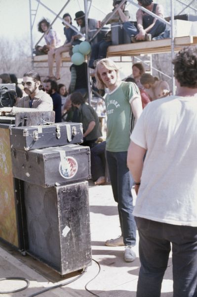 Grateful Dead drummer Mickey Hart stands behind equipment to the left of the band's road manager, Jon McIntyre, who is wearing a t-shirt that reads "Fillmore East," while people can be seen milling about behind them and sitting on scaffolding.