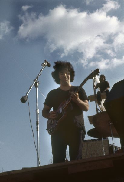 Jerry Garcia, guitarist for the Grateful Dead, playing on stage at the Sound Storm festival. An audience member is visible sitting on the scaffolding behind the stage.