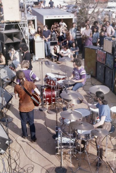 Elevated view of the Grateful Dead mid-performance as viewed from scaffolding above the stage, in front of rows of speakers and amplifiers. Audience members are seen in the wings of the stage.