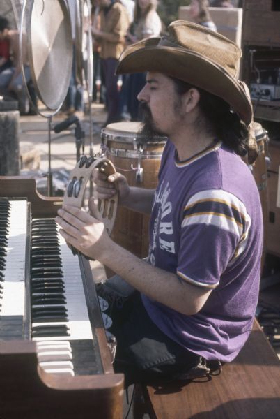 Ron "Pigpen" McKernan, keyboardist and percussionist for the Grateful Dead, sitting at his keyboard mid-performance, playing a tambourine. His t-shirt reads "Fillmore East."