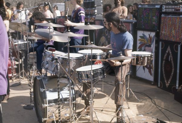 Side view from stage of Bill Kreutzmann and Mickey Hart playing drums on stage with the Grateful Dead while Phil Lesh stands between them. Behind the band are multi-colored amplifiers and speakers, as well as a few audience members.