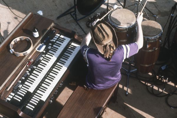 View from above of Ron "Pigpen" McKernan, keyboardist and percussionist for the Grateful Dead, sitting at his keyboard mid-performance playing conga drums with mallets. A bottle of Old Style beer sits on the keyboard next to a tambourine.