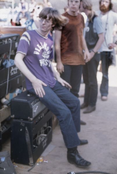 Phil Lesh, bassist for the Grateful Dead, sits on a small amplifier backstage, while Bob Weir stands behind him.