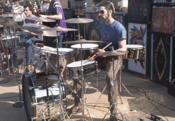 Bill Kreutzmann and Mickey Hart playing drums on stage with the Grateful Dead. Phil Lesh, the bassist, is between them. In the background are multi-colored amplifiers and speakers, as well as a few audience members.