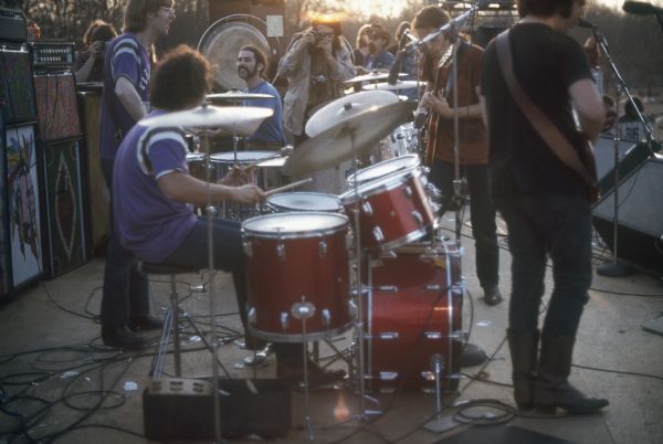 The Grateful Dead featuring Phil Lesh, Mickey Hart, Bill Kreutzmann, Bob Weir, and Jerry Garcia, performing on stage with the sun setting behind them. Ron "Pigpen" McKernan is in the center background, and people are taking photographs from the wings of the stage.