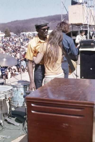 The drummer for Luther Allison being embraced by a woman on stage behind his drum kit. Behind them the bassist is visible, and the crowd in front of the stage is visible in the background.