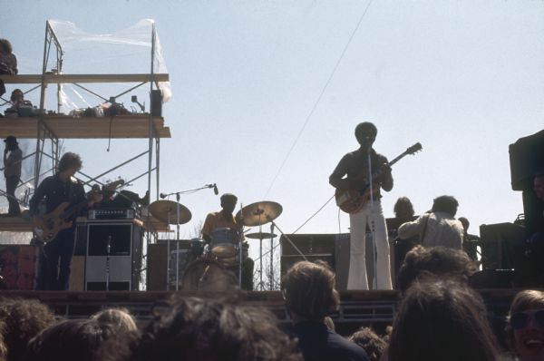 View from audience of guitarist Luther Allison with his band, which is made up of a drummer and bassist. A photographer is crouched in front of Allison. People are standing and sitting on scaffolding which is visible in the left background.