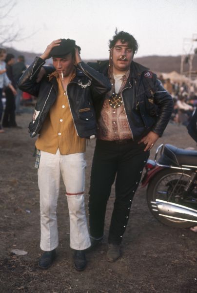 Two bikers at the Sound Storm music festival. The biker on the left is wearing a hat, leather jacket, white pants, and is smoking a cigarette. The biker on the right has a large ring through his nose, a headband over his hair. He is also wearing a large swastika necklace and leather jacket.