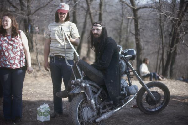 A biker wearing a long dark coat, a headband, and sunglasses sits on a motorcycle with an unusual metal backrest. A man and a woman are standing next to him, and more people are in the woods in the background.