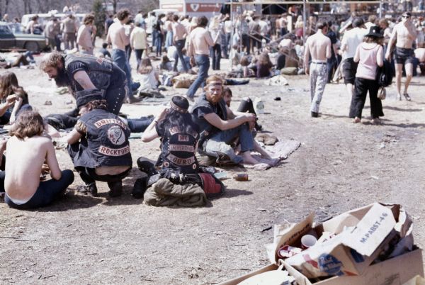 A group of bikers wearing leather jackets representing their gang affiliations sit and stand on the ground. In the background is a crowd of people near the Sound Storm stage and scaffolding. Some of the biker's jackets read: "Drifters MC Rockford" and "Drifters MC Property of Guru."