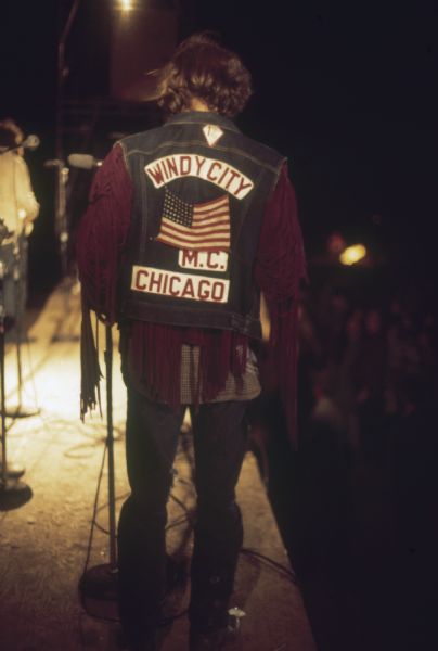 A biker wearing a fringed leather jacket with a sleeveless denim jacket over it that reads "Windy City M.C. Chicago" stands on the side of the Sound Storm stage at night.