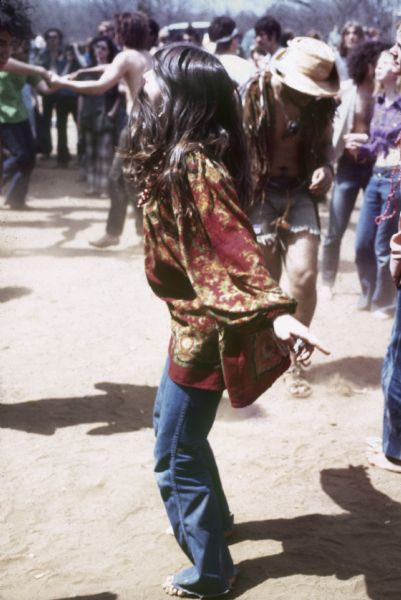 A young woman wearing a patterned blouse and blue jeans dances among a crowd of other audience members.