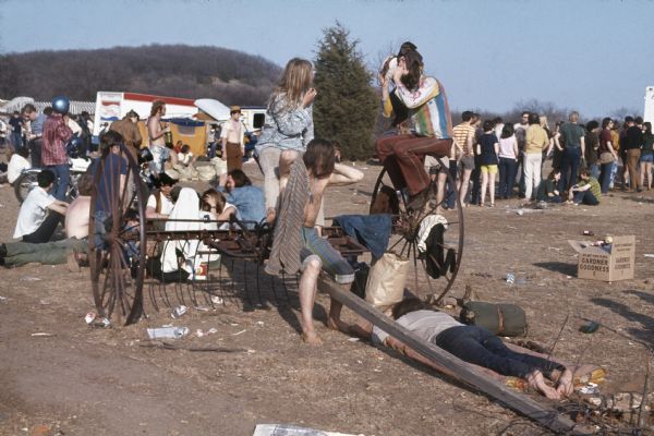 A group of audience members crowd onto an old cultivator near the Sound Storm stage. A line has formed behind them to the right, and there is a man with a motorcycle to the left. Beer cans and other garbage can be seen on the ground around them. In the background are trucks, vans, and tents.