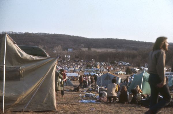 The Sound Storm music festival site as seen from the camping area. Attendees mill around many tents, campers and automobiles. In the far background a large crowd is in front of the stage.