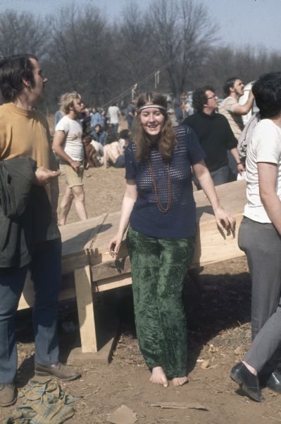 A barefoot young female audience member wearing a macrame shirt and smoking a cigarette leans against the ramp to the main stage at the Sound Storm music festival with a crowd of people visible around her.