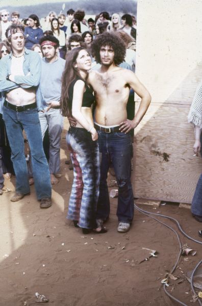 A young couple poses at the foot of the Sound Storm stage with audience members crowding behind them. The young woman is wearing tie-dyed pants and the young man is shirtless.