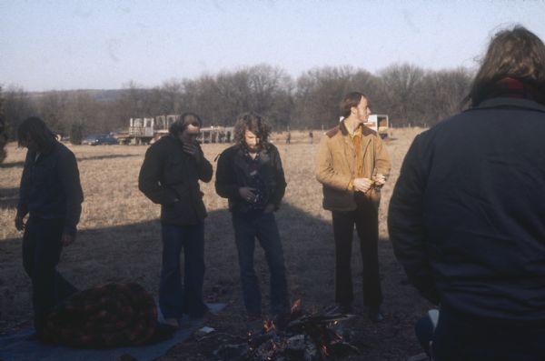 A group of young men crowd around a campfire in the Sound Stage campground. In the background is a truck and a station wagon near men who appear to be setting up the stage a day or two before the audience will arrive.