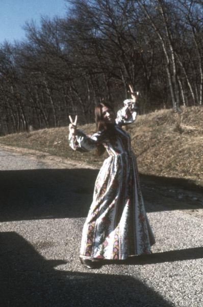 A smiling woman in a long pattered dress stands in the road as crowded cars slowly roll past, making a peace sign with her hands for a captive audience.