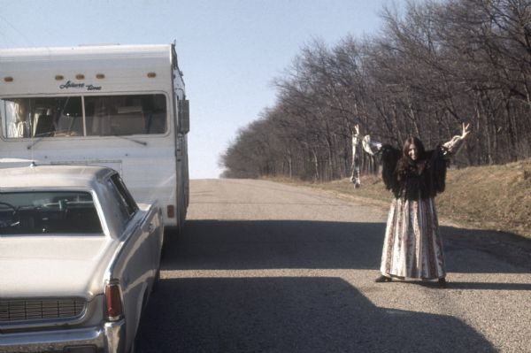 A smiling woman in a long pattered dress stands in the road next to a crowded camper, making a peace sign with her hands for a captive audience.