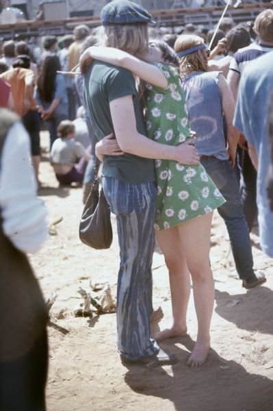 A young couple kisses in the audience of the Sound Storm music festival. The young woman is wearing a flower print dress and the young man is wearing a hat with tie-dyed pants. In the background is a crowd in front of the stage.