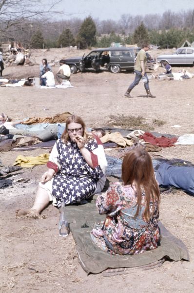 Two young female audience members, one wearing a paisley print dress, the other wearing an American flag print dress, sit in the camping area smoking marijuana. A small crowd of other campers sits in the background.
