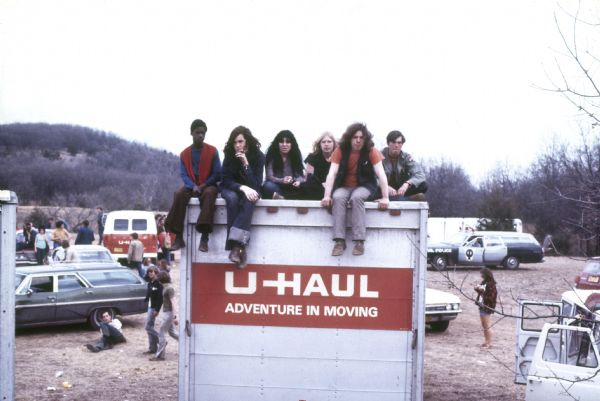 Six young people crowd on top of the rear roof of a U-Haul trailer in the camping area of the Sound Storm music festival. There are other audience members with cars and trailers in the background.