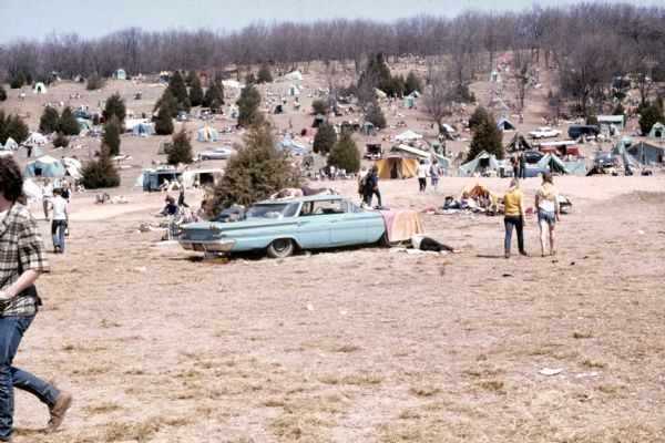 A wide view of the camping area at the site of the Sound Storm music festival. People are walking among tents and vehicles spread out on a hill. There is a car parked in the foreground with people sleeping in and around it.