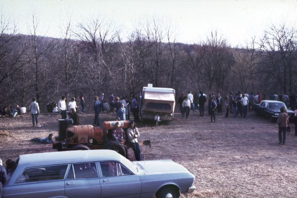 A group of audience members crowd around a camper, and two automobiles. Two men are leaning on a trailer carrying large tanks, perhaps generators.