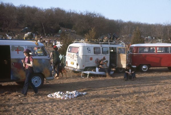 Camping site with Volkswagen vans and tents. People are milling around the camping area of the Sound Storm music festival. "Speed Kills" is painted on the back of one of the vans. More tents are on the hill in the background.