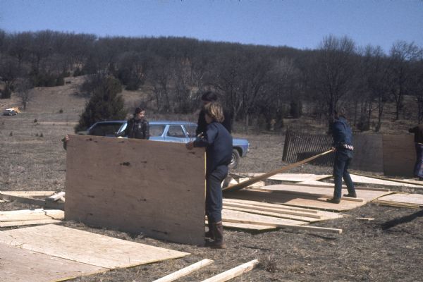 A group of three young workers amid construction of plywood stalls. There is a station wagon in the background. A few tents are set up on the hill in the background.