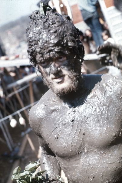 A bearded young man, Michael McCormick of Madison, was crowned "The Mud King." He is covered in dark mud from the banks of nearby Rowan Creek and holding a plant while standing in the audience at the Sound Storm music festival.