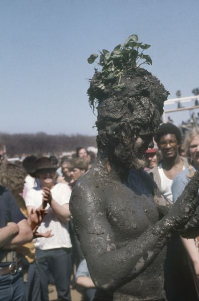 A bearded young man, Michael McCormick of Madison, was crowned "The Mud King." He is covered in dark mud from the banks of nearby Rowan Creek and is standing in the audience at the Sound Storm music festival with a plant embedded in the pile of mud on his head.