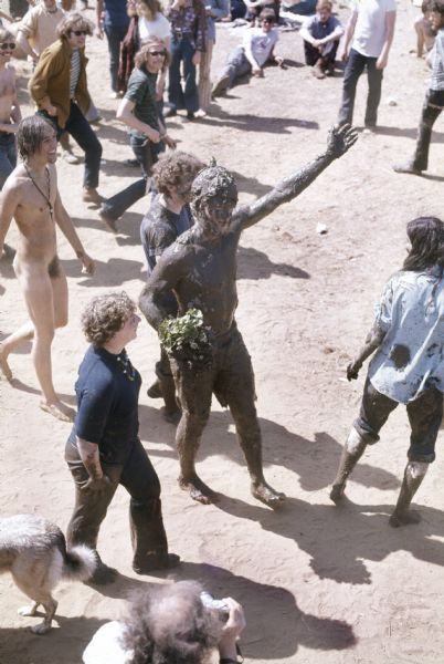 A bearded young man, Michael McCormick of Madison, was crowned the "The Mud King." He is covered in dark mud from the banks of nearby Rowan Creek holding a plant while walking through the audience at the Sound Storm music festival. Behind him a naked man and others with mud on their clothes are following him. In the foreground is a man with a camera.