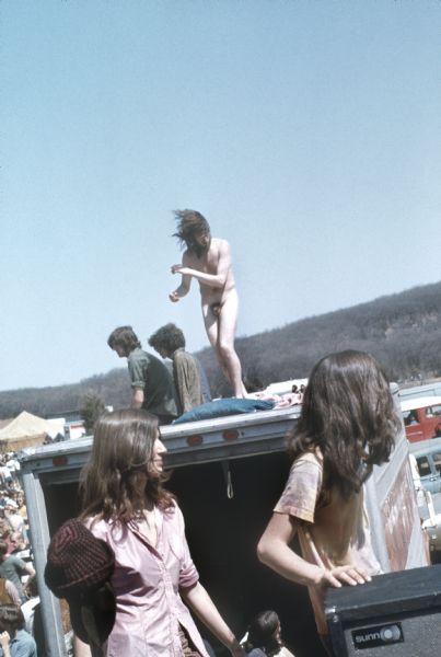 A nude man with long hair dances on top of a U-Haul moving truck. Two people in the foreground are near a Sunn brand amplifier on the bottom right.